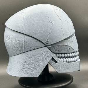 May include: A white, 3D printed helmet with a textured surface and a detailed, toothy grin. The helmet is designed with a segmented, articulated jaw.