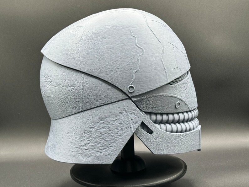 May include: A white, 3D printed helmet with a textured surface and a detailed, toothy grin. The helmet is designed with a segmented, articulated jaw.