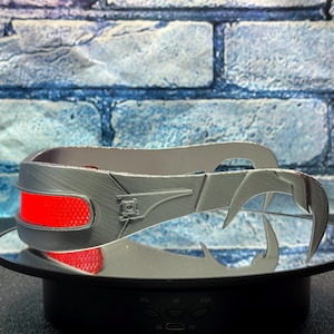 May include: A silver-colored, claw-like headpiece with a red accent. The headpiece is displayed on a mirrored surface against a blue brick wall background. The headpiece has a textured surface and a futuristic design.