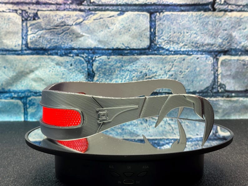 May include: A silver-colored, claw-like headpiece with a red accent. The headpiece is displayed on a mirrored surface against a blue brick wall background. The headpiece has a textured surface and a futuristic design.