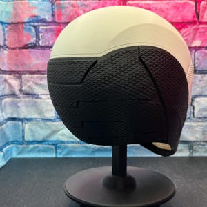 May include: A two-tone helmet, with a cream-colored top and a black textured lower section. The helmet is mounted on a black stand, set against a brick wall backdrop.