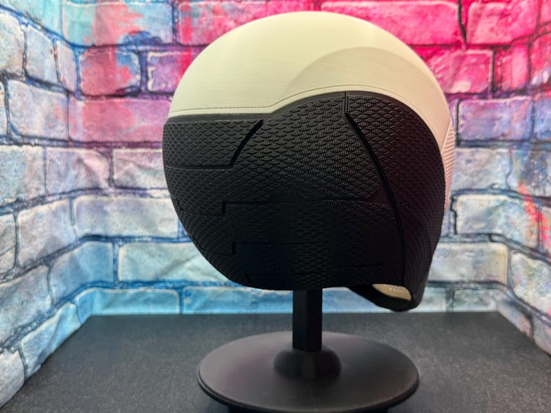 May include: A two-tone helmet, with a cream-colored top and a black textured lower section. The helmet is mounted on a black stand, set against a brick wall backdrop.