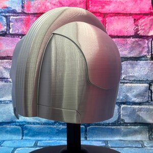 May include: A silver-colored helmet with a textured surface, displayed on a black stand. The helmet has a rounded top and a curved visor-like section. The background is a brick wall with blue, pink, and purple paint.
