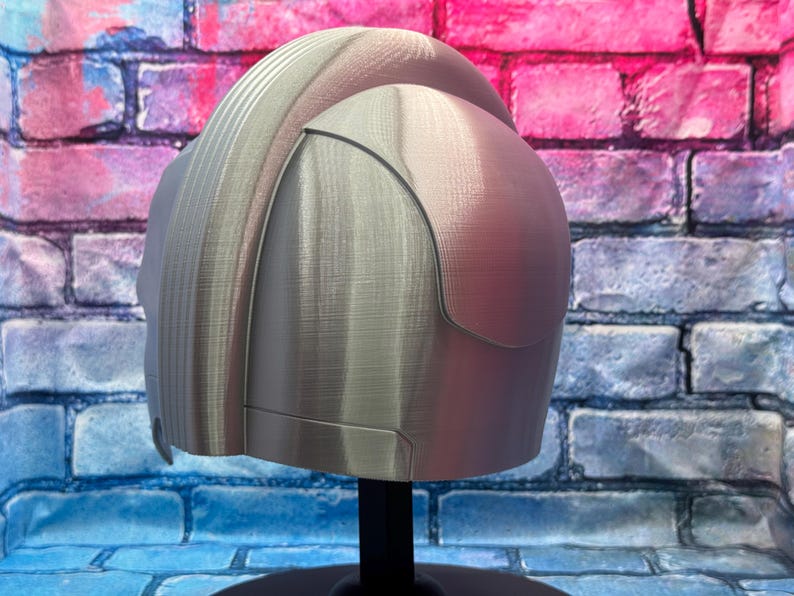 May include: A silver-colored helmet with a textured surface, displayed on a black stand. The helmet has a rounded top and a curved visor-like section. The background is a brick wall with blue, pink, and purple paint.