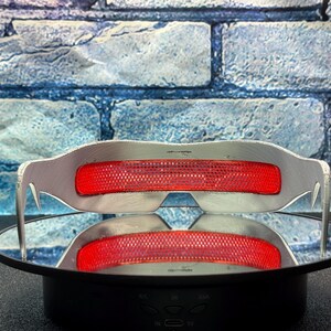 May include: Silver-framed glasses with a bright red, mesh-like center section. The glasses are displayed on a reflective surface, with a brick-patterned background. The glasses have a modern, futuristic design.