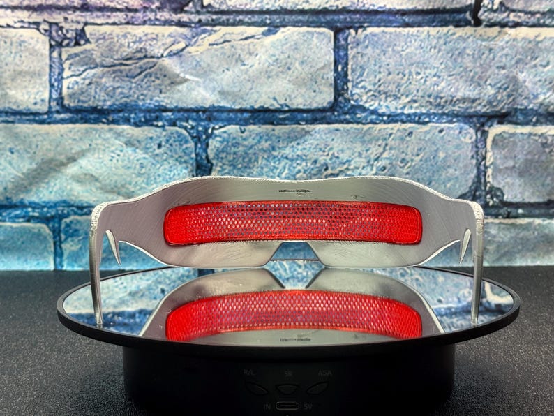 May include: Silver-framed glasses with a bright red, mesh-like center section. The glasses are displayed on a reflective surface, with a brick-patterned background. The glasses have a modern, futuristic design.