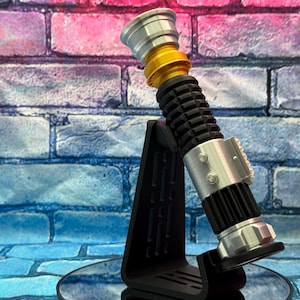 May include: A lightsaber prop in silver, gold, and black, presented on a black stand. The hilt has a ribbed black grip, silver details, and a gold section. The prop is set against a brick wall with pink and blue tones.