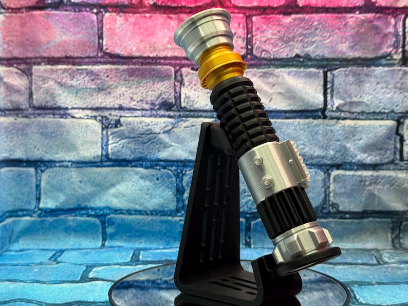 May include: A lightsaber prop in silver, gold, and black, presented on a black stand. The hilt has a ribbed black grip, silver details, and a gold section. The prop is set against a brick wall with pink and blue tones.
