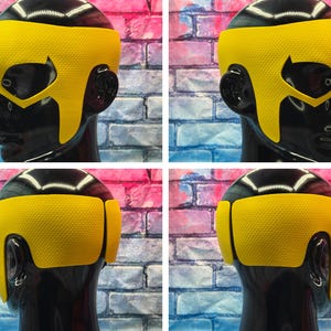 K&ouml;nnte beinhalten: Eine leuchtend gelbe, texturierte Superheldenmaske auf einem schwarzen Schaufensterpuppenkopf. Die Maske hat ein breites Stirnband, spitze Augenausschnitte und Seitenteile, die bis zum Kiefer reichen und das Gesicht sowie die Ohren bedecken.