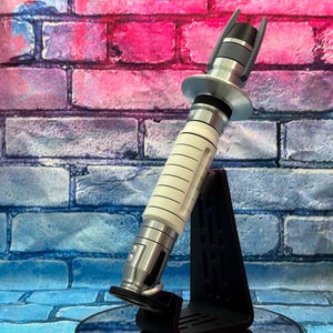 May include: A futuristic lightsaber prop with a white and silver handle, black accents, and a silver emitter. The prop is displayed on a black stand against a brick wall backdrop with pink and blue hues.