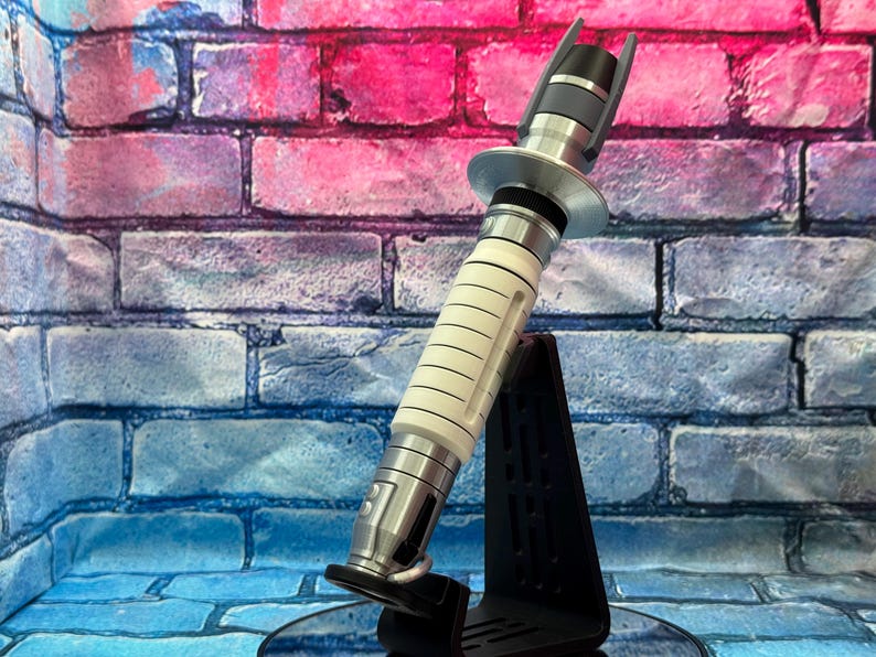 May include: A futuristic lightsaber prop with a white and silver handle, black accents, and a silver emitter. The prop is displayed on a black stand against a brick wall backdrop with pink and blue hues.