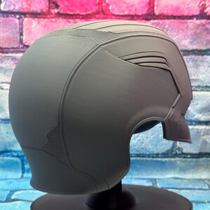 May include: A gray helmet with a smooth, matte finish. The helmet has a streamlined design with textured details on the top and sides. It is displayed on a black stand against a colorful brick wall.