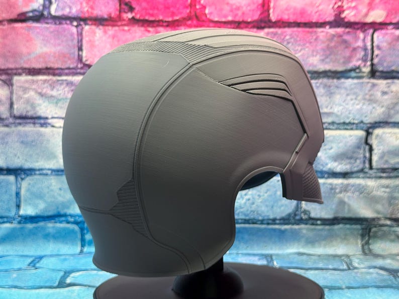 May include: A gray helmet with a smooth, matte finish. The helmet has a streamlined design with textured details on the top and sides. It is displayed on a black stand against a colorful brick wall.