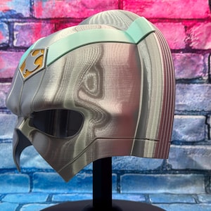 May include: A silver and teal colored helmet with a unique design. The helmet has a visor, a pointed chin, and a gold emblem on the side. The helmet is displayed on a black stand against a brick wall.