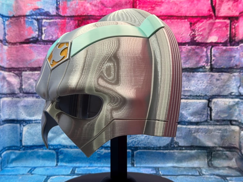 May include: A silver and teal colored helmet with a unique design. The helmet has a visor, a pointed chin, and a gold emblem on the side. The helmet is displayed on a black stand against a brick wall.