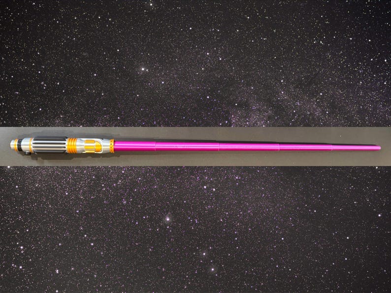 May include: A pink lightsaber with a silver, black, and gold hilt. The lightsaber is displayed horizontally against a starry background. The hilt features intricate details and a metallic finish. The blade is a vibrant pink color.