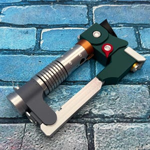 May include: A futuristic-looking device with a silver, ribbed cylindrical body, a dark green angular component, and a silver blade. The device has a copper-colored ring and a red accent. The background is a blue brick wall.