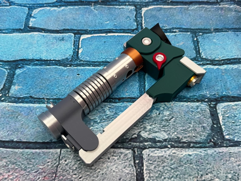 May include: A futuristic-looking device with a silver, ribbed cylindrical body, a dark green angular component, and a silver blade. The device has a copper-colored ring and a red accent. The background is a blue brick wall.