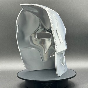 May include: A white 3D printed mask with a textured surface and a detailed design. The mask has a large eye opening and a smaller opening for the mouth.