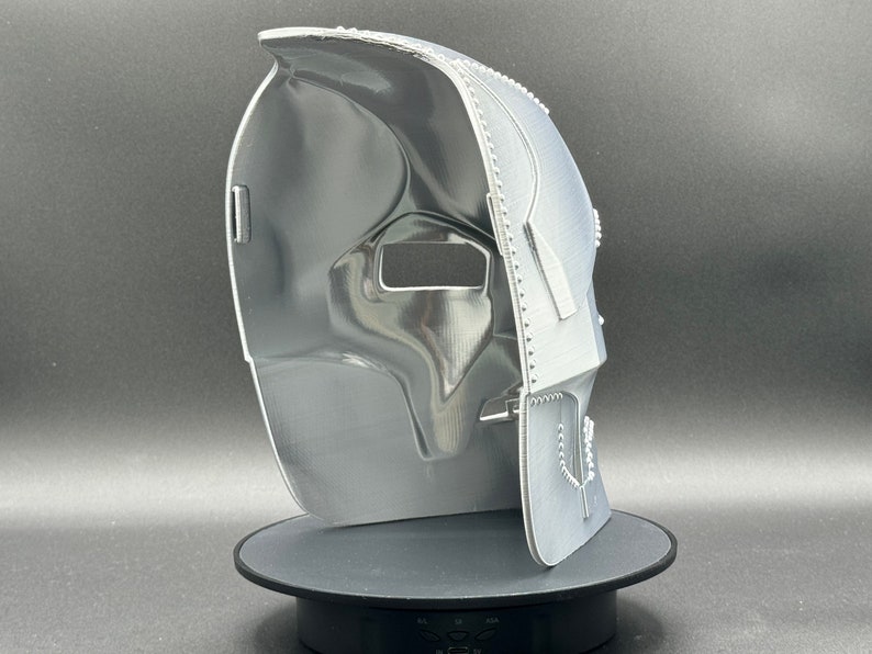 May include: A white 3D printed mask with a textured surface and a detailed design. The mask has a large eye opening and a smaller opening for the mouth.