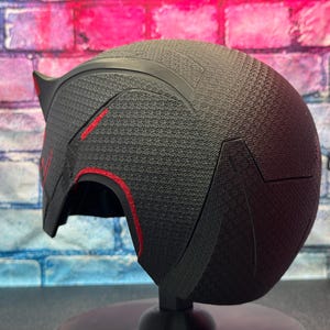 May include: A black helmet with red accents. The helmet has a textured surface and a pointed design on the top. The red accents are visible along the edges and on the front of the helmet. The helmet is displayed on a black stand.