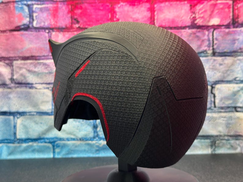 May include: A black helmet with red accents. The helmet has a textured surface and a pointed design on the top. The red accents are visible along the edges and on the front of the helmet. The helmet is displayed on a black stand.