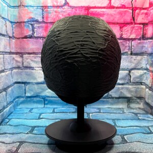 May include: A black, textured sphere on a black stand. The sphere has a rough, organic texture. The stand has a circular base and a slender stem. The background is a brick wall with blue, pink, and purple hues.
