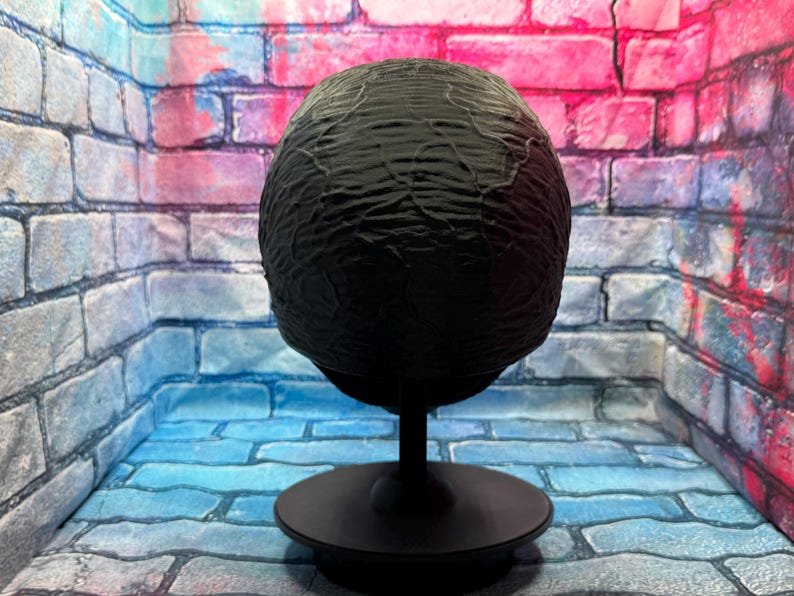 May include: A black, textured sphere on a black stand. The sphere has a rough, organic texture. The stand has a circular base and a slender stem. The background is a brick wall with blue, pink, and purple hues.
