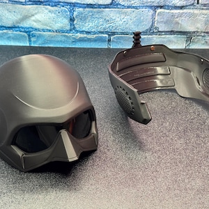 May include: A matte black helmet with integrated tinted visor and a separate face guard. The helmet has a smooth, rounded design with angular features around the visor. The face guard has a speaker and a mounting mechanism.