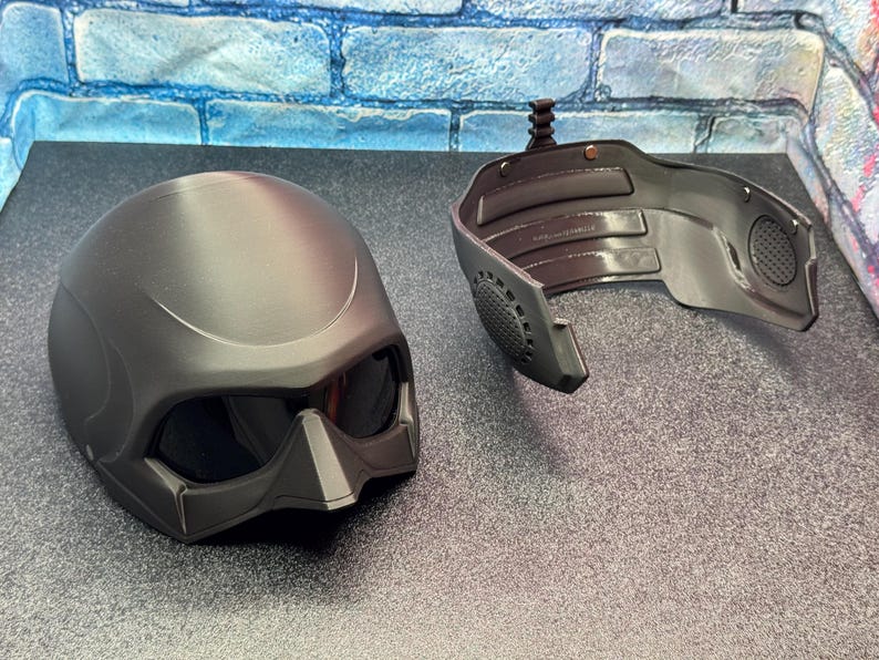 May include: A matte black helmet with integrated tinted visor and a separate face guard. The helmet has a smooth, rounded design with angular features around the visor. The face guard has a speaker and a mounting mechanism.
