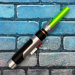May include: A toy lightsaber with a green blade and a silver and black handle. The handle has a red button and gold accents. The lightsaber is resting on a blue brick background.