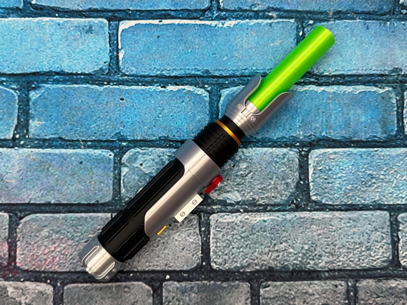 May include: A toy lightsaber with a green blade and a silver and black handle. The handle has a red button and gold accents. The lightsaber is resting on a blue brick background.