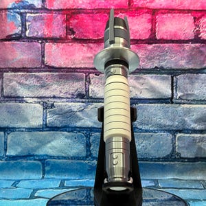 May include: A silver and white lightsaber prop, featuring a cylindrical handle with horizontal grooves and a metallic top and bottom. The prop is displayed on a black stand against a brick wall background.