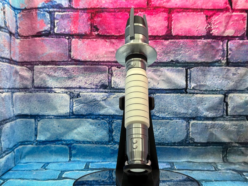 May include: A silver and white lightsaber prop, featuring a cylindrical handle with horizontal grooves and a metallic top and bottom. The prop is displayed on a black stand against a brick wall background.