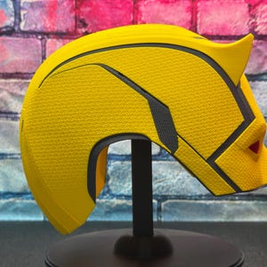 Puede incluir: Un casco de superh&eacute;roe amarillo con detalles en negro y gris, exhibido en un soporte negro. El casco tiene una superficie texturizada y un dise&ntilde;o puntiagudo en los lados. El fondo presenta una pared de ladrillos.