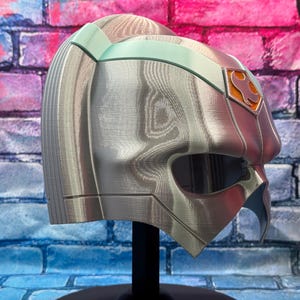 May include: A silver and teal-accented helmet with an orange emblem, displayed on a black stand. The helmet has a detailed, textured surface and a visor opening. The background is a colorful brick wall.