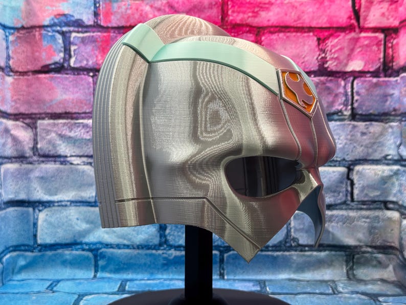 May include: A silver and teal-accented helmet with an orange emblem, displayed on a black stand. The helmet has a detailed, textured surface and a visor opening. The background is a colorful brick wall.