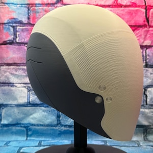 May include: A futuristic-looking helmet with a two-tone design. The top is a light beige, while the lower section is dark gray. The helmet has a textured surface and is mounted on a black stand. Two silver rivets are visible on the side.