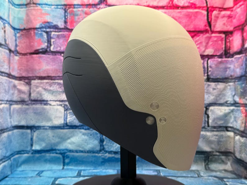 May include: A futuristic-looking helmet with a two-tone design. The top is a light beige, while the lower section is dark gray. The helmet has a textured surface and is mounted on a black stand. Two silver rivets are visible on the side.