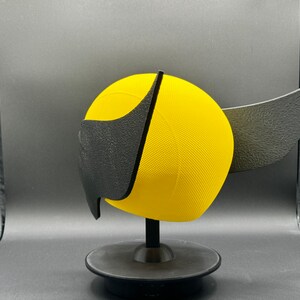 Puede incluir: Un casco amarillo y negro impreso en 3D con un ala en el lateral. El casco est&aacute; en un soporte negro.