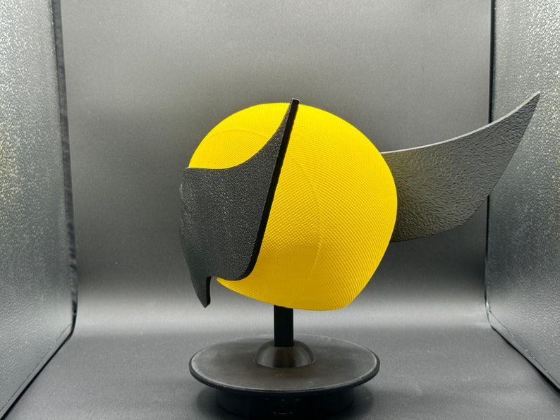 Puede incluir: Un casco amarillo y negro impreso en 3D con un ala en el lateral. El casco est&aacute; en un soporte negro.