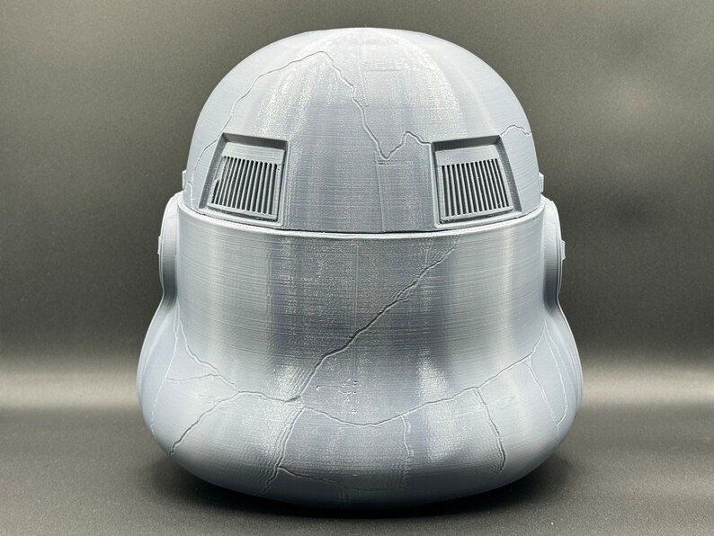 May include: A gray plastic helmet with a textured surface and two rectangular vents on the top. The helmet has a cracked appearance.