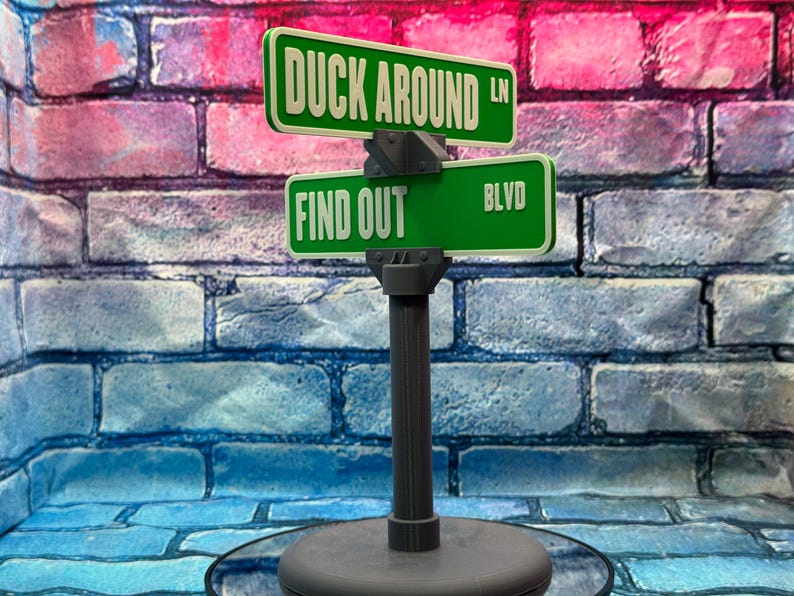 May include: A novelty street sign with green signs that read "DUCK AROUND LN" and "FIND OUT BLVD". The signs are mounted on a gray post and base, set against a brick wall background.