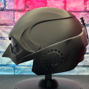 May include: A matte black helmet with a futuristic design, featuring a visor, side vents, and a textured surface. The helmet is displayed on a black stand, showcasing its sleek, angular form. This item is likely a cosplay or collectible piece.