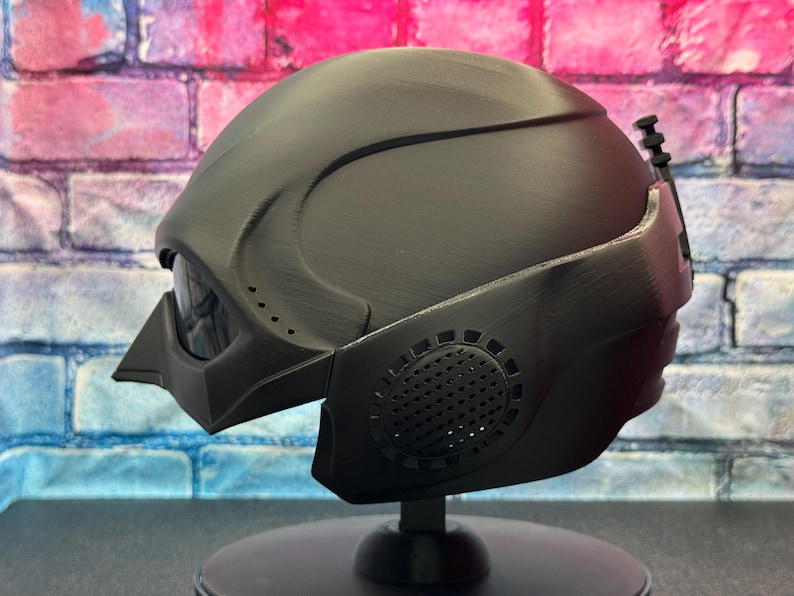 May include: A matte black helmet with a futuristic design, featuring a visor, side vents, and a textured surface. The helmet is displayed on a black stand, showcasing its sleek, angular form. This item is likely a cosplay or collectible piece.