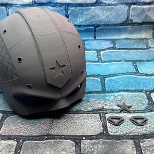 May include: A gray, 3D-printed helmet with a star emblem and diamond-patterned side panel. Includes a star-shaped accessory and two small, triangular pieces. The helmet has several small circular openings and a visor area.