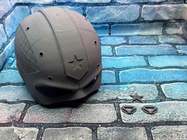 May include: A gray, 3D-printed helmet with a star emblem and diamond-patterned side panel. Includes a star-shaped accessory and two small, triangular pieces. The helmet has several small circular openings and a visor area.