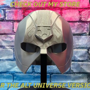 May include: A silver and red superhero mask with a central emblem, displayed on a black stand. The mask has eye openings and is set against a brick wall with text that reads "CHECK OUT MY STORE" and "FOR THE ALT UNIVERSE VERSION!"