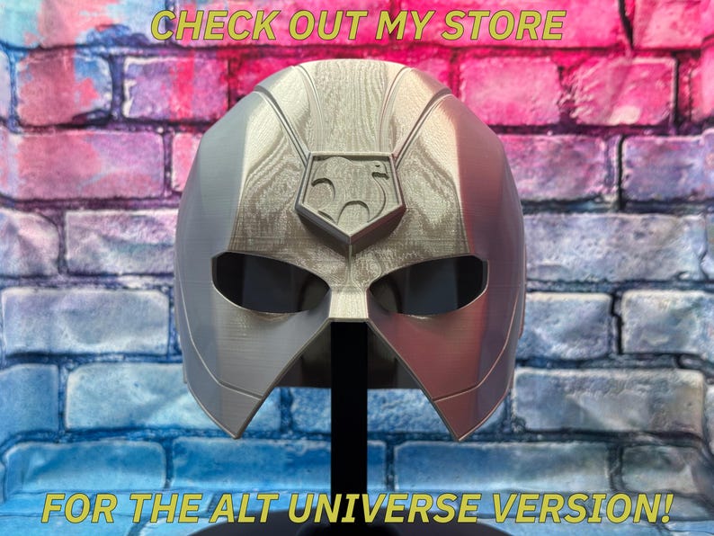 May include: A silver and red superhero mask with a central emblem, displayed on a black stand. The mask has eye openings and is set against a brick wall with text that reads "CHECK OUT MY STORE" and "FOR THE ALT UNIVERSE VERSION!"