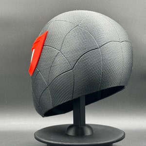 Puede incluir: Una m&aacute;scara impresa en 3D de color negro y rojo con un dise&ntilde;o de telara&ntilde;a. La m&aacute;scara est&aacute; en un soporte negro.